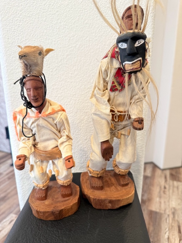 Two carved dancer figures in cream clothing, one wearing a deer headdress and the other a dark mask.