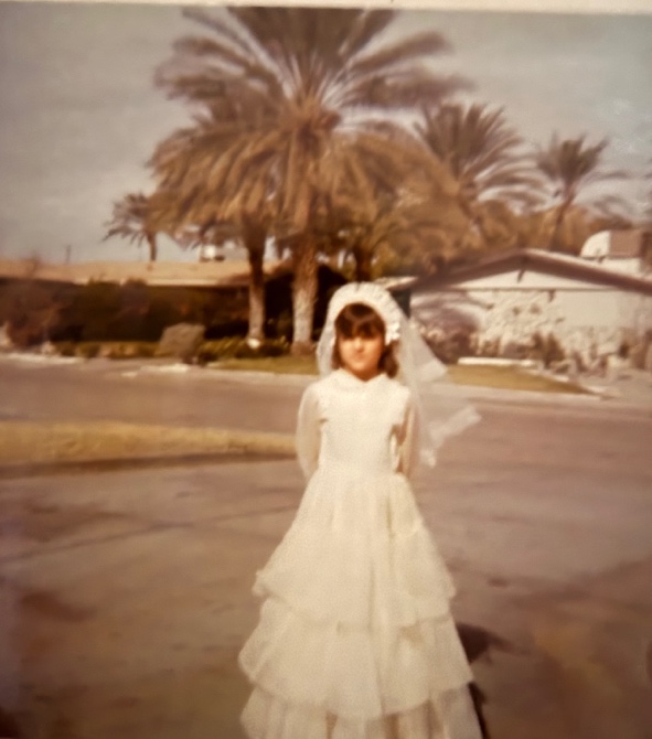 A child in a white Communion dress and veil standing on a residential street lined with palm trees.
