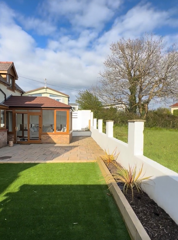 A sunlit back garden with a conservatory, lawn, white wall and large tree beyond it.